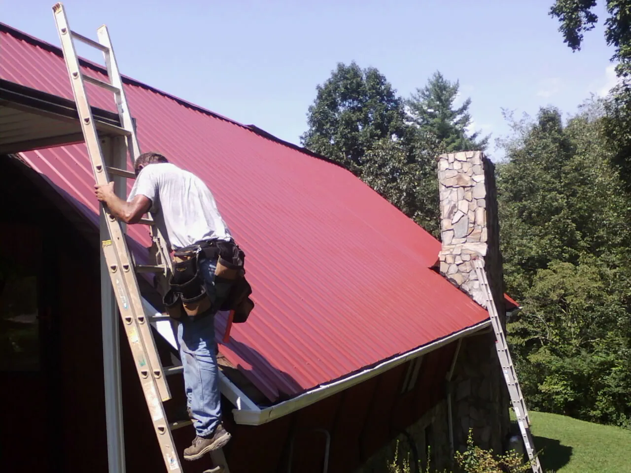 Roofer on a ladder beside a red metal roof and stone chimney — Soffit & Fascia Repair by Roofing Brothers in Chester Heights